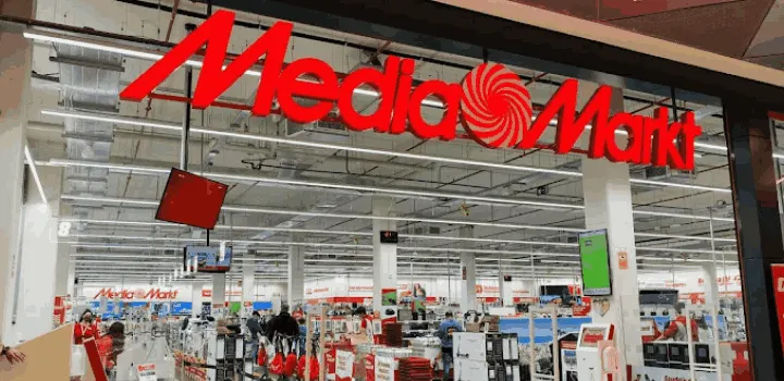 Letrero rojo "Media Markt" en la entrada de una tienda grande. Dentro hay estanterías llenas de productos y gente comprando.