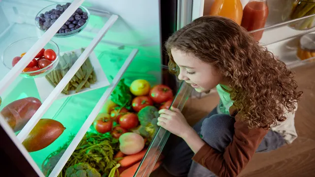 Imagen de una niña abriendo el cajon de un frigorífico lleno de frutas y verduras frescas.