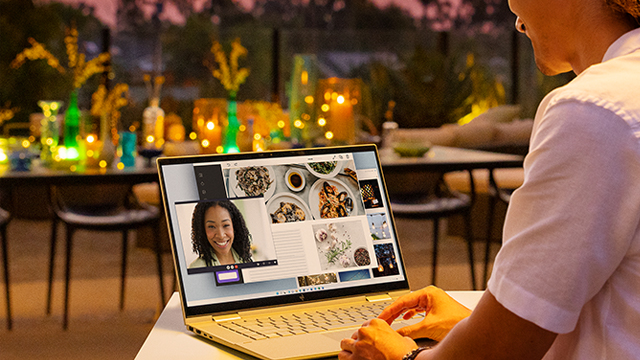 Persona usa un portátil con videollamada en un patio con mesas, sillas y luces decorativas.