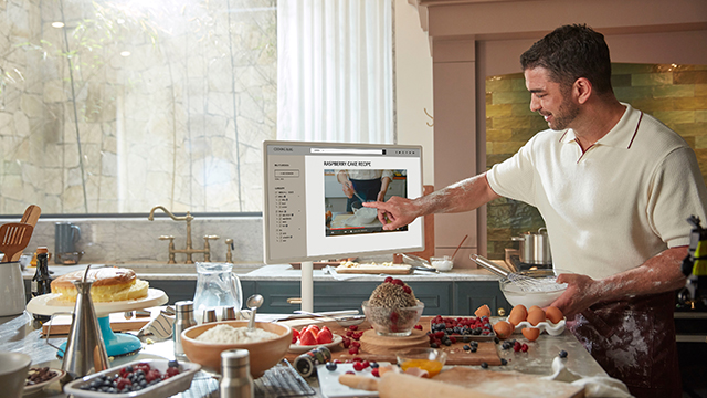 Un hombre en una cocina apunta a un monitor que muestra una receta, rodeado de ingredientes para hornear como harina, huevos y fruta.
