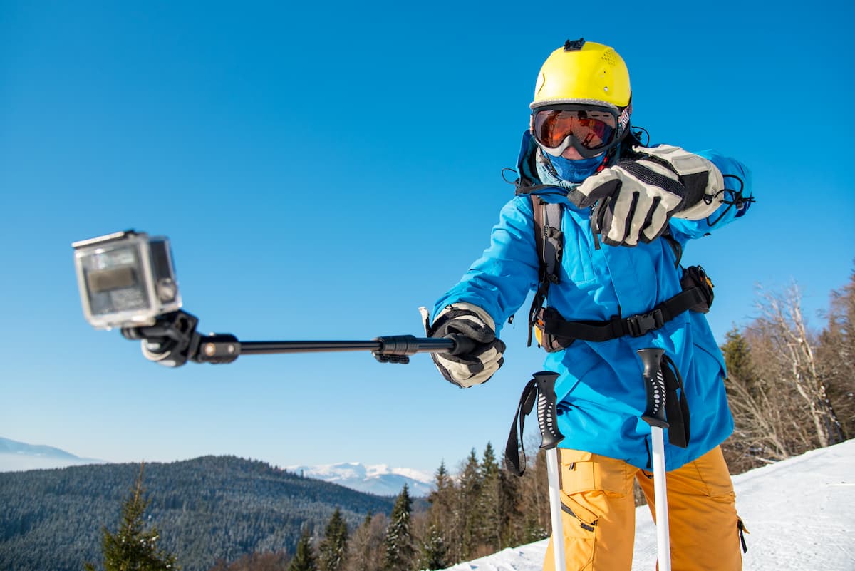 Esquiador con casco amarillo grabándose con una GoPro en palo selfie en montaña nevada.