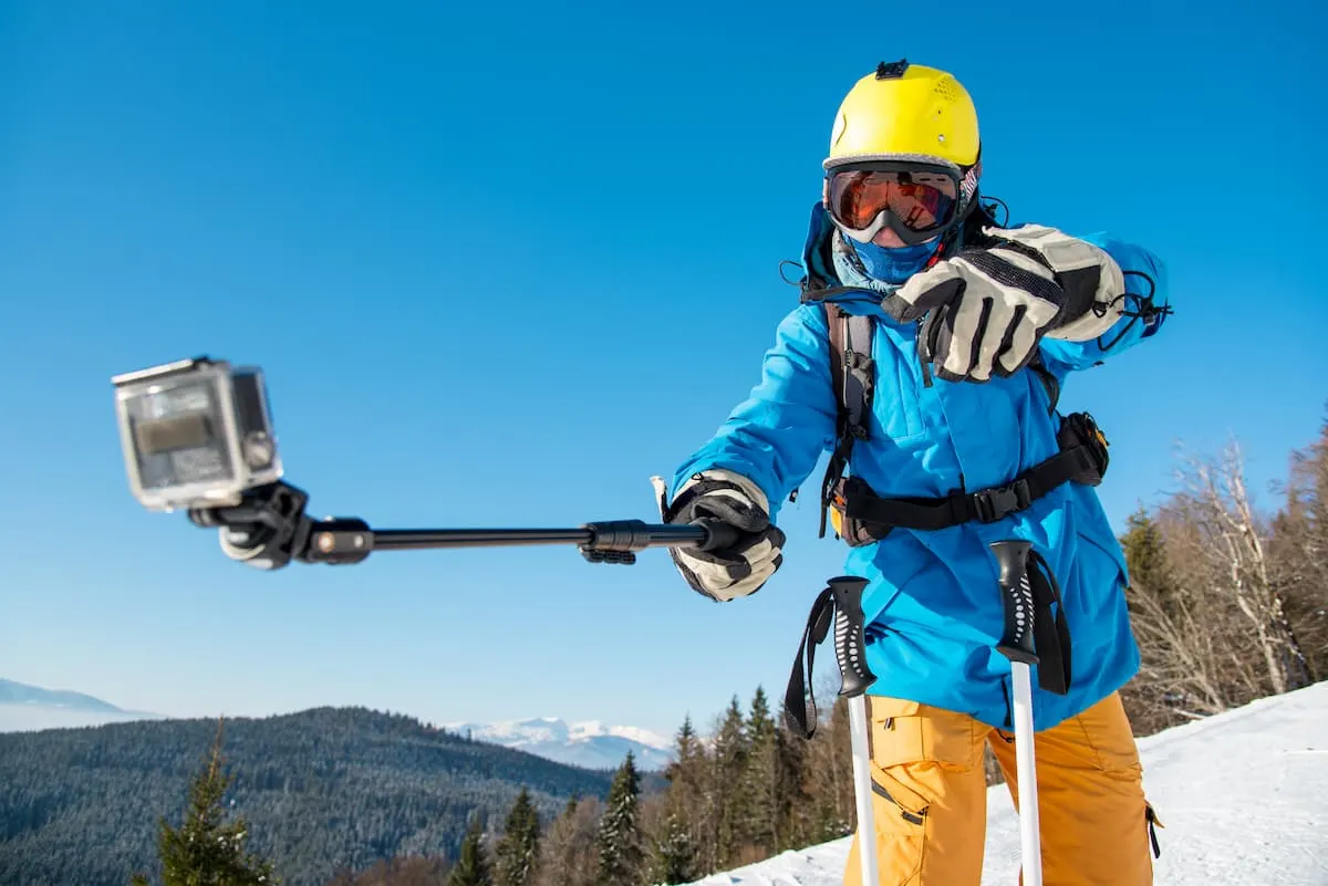 Esquiador con casco amarillo grabándose con una GoPro en palo selfie en montaña nevada.