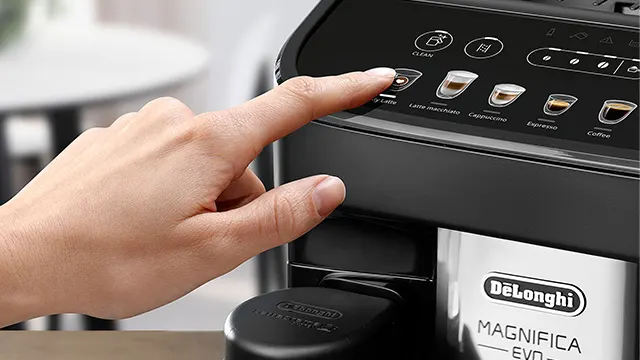 Una mano presiona el botón de inicio en una cafetera automática negra y plateada DeLonghi Magnifica Evo. Los botones tienen iconos de café.