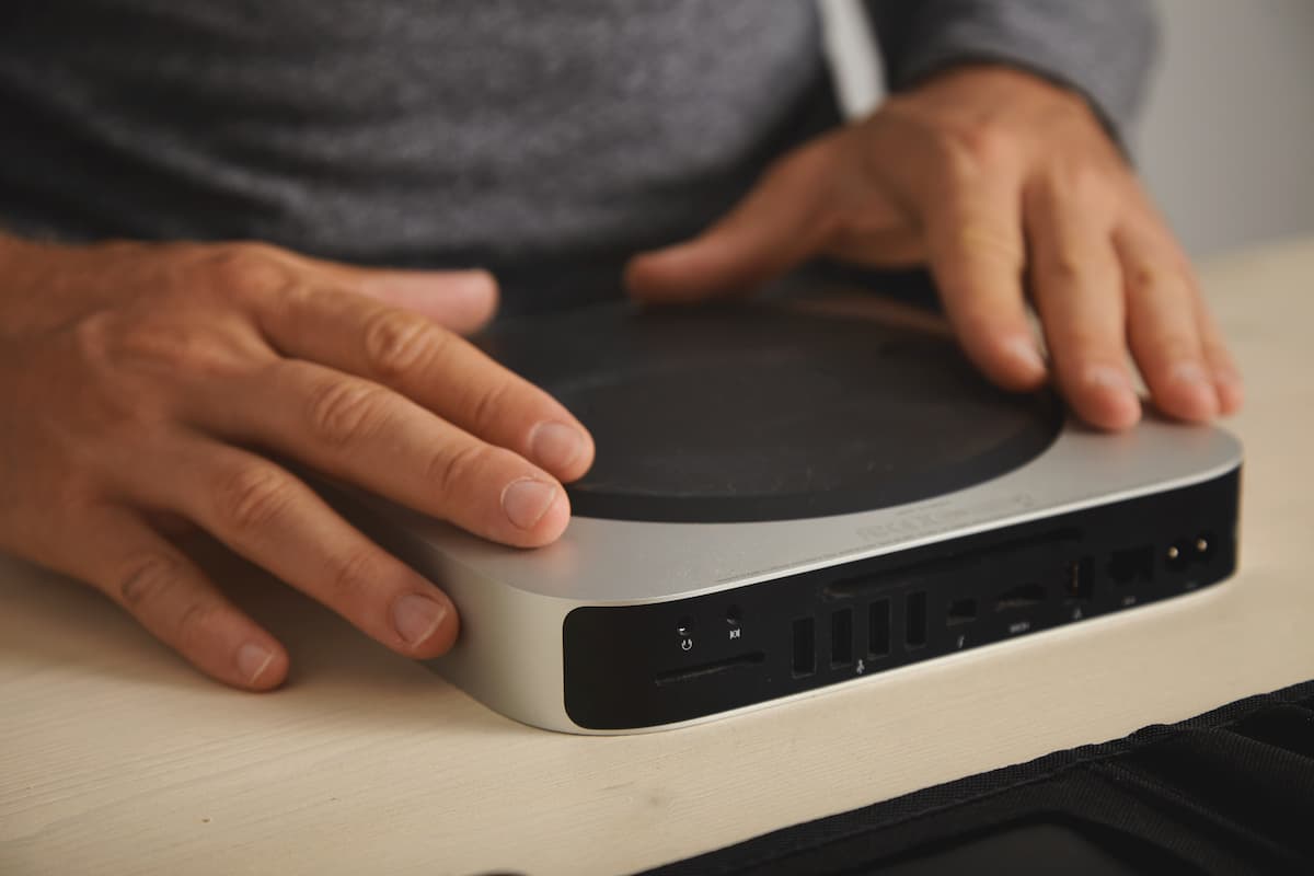 Manos colocando un mini PC plateado sobre un escritorio, mostrando los puertos traseros del equipo.