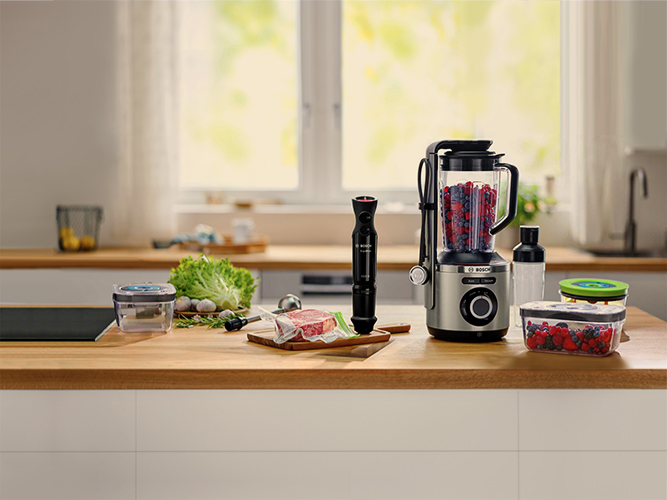 En una cocina, hay una batidora, una batidora de inmersión, lechuga, un filete, y recipientes con bayas sobre una encimera de madera.