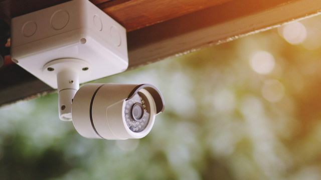 Cámara de seguridad blanca montada en techo de madera. Fondo borroso con vegetación verde y tonos dorados de luz solar.
