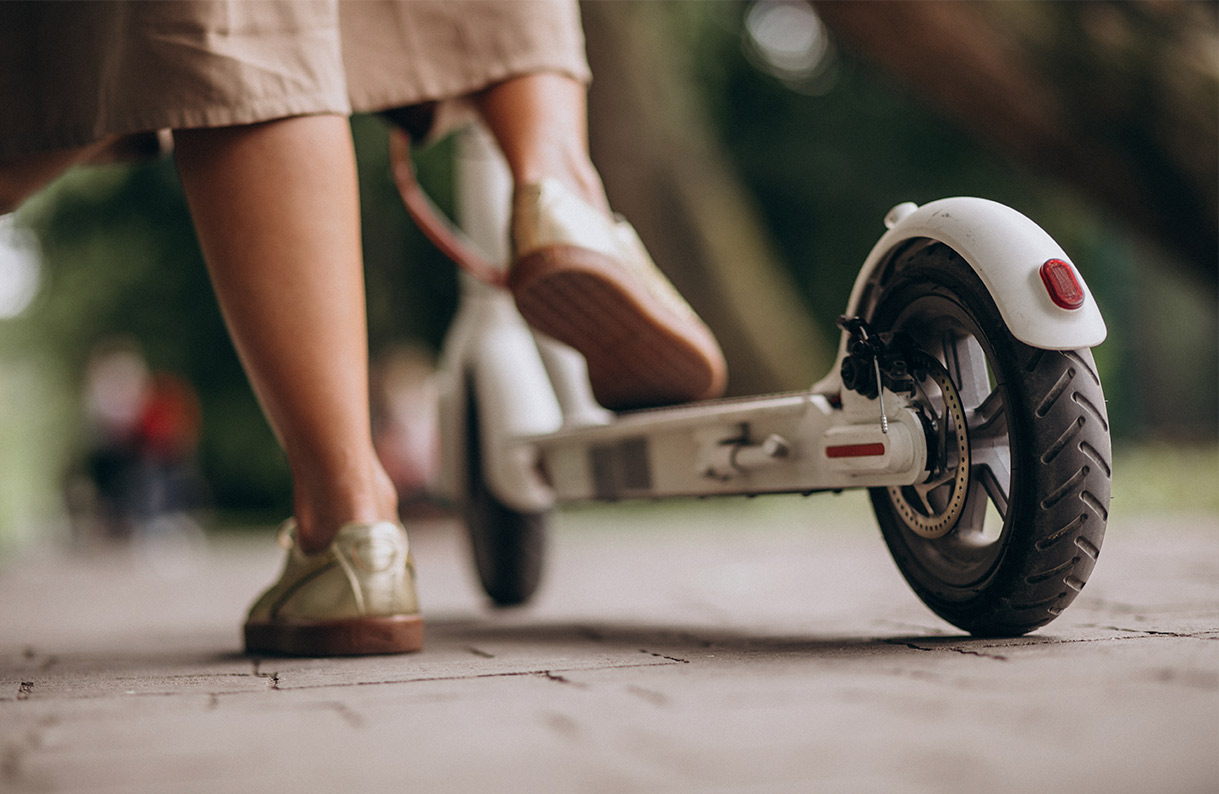 Vista cercana de una persona en un patinete blanco con zapatos dorados sobre un pavimento de baldosas. El fondo es borroso y verde.