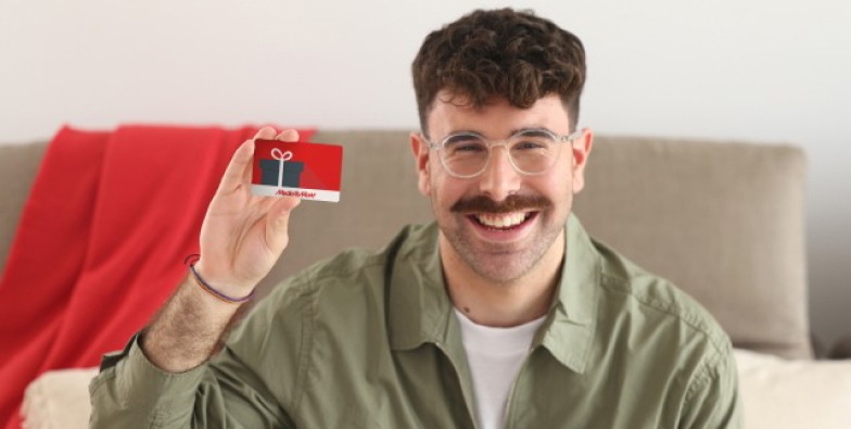 Un hombre sonriente sostiene una tarjeta roja con un regalo negro. Lleva una camisa verde, gafas y está sentado en un sofá.