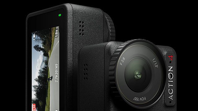 Cámara de acción negra con lente y pantalla LCD mostrando un camino rural bordeado de árboles. Una luz verde indica el estado.