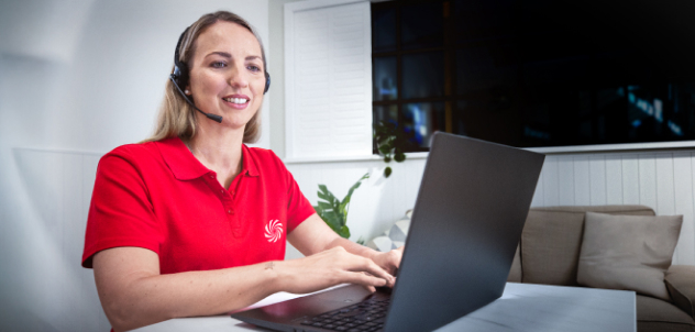 Mujer con auriculares y polo rojo trabaja en un portátil, atendiendo a clientes desde una sala de estar luminosa.