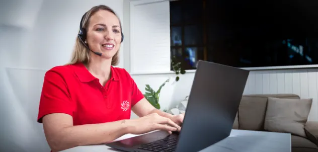 Mujer con auriculares y polo rojo trabaja en un portátil, atendiendo a clientes desde una sala de estar luminosa.