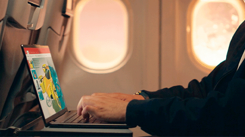 En un avión, una persona usa una computadora portátil. Se ven ventanas de avión con luz exterior. El asiento está tapizado.