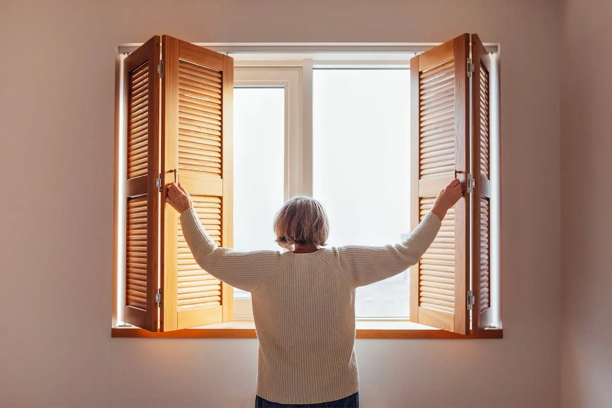 Persona mayor abriendo las contraventanas de madera de una ventana, de espaldas, dejando entrar la luz natural en la habitación.