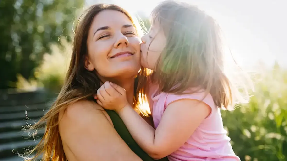 Una niña abraza a una mujer con camisa verde. La niña usa una camisa rosa. Hay hierba y árboles en el fondo.