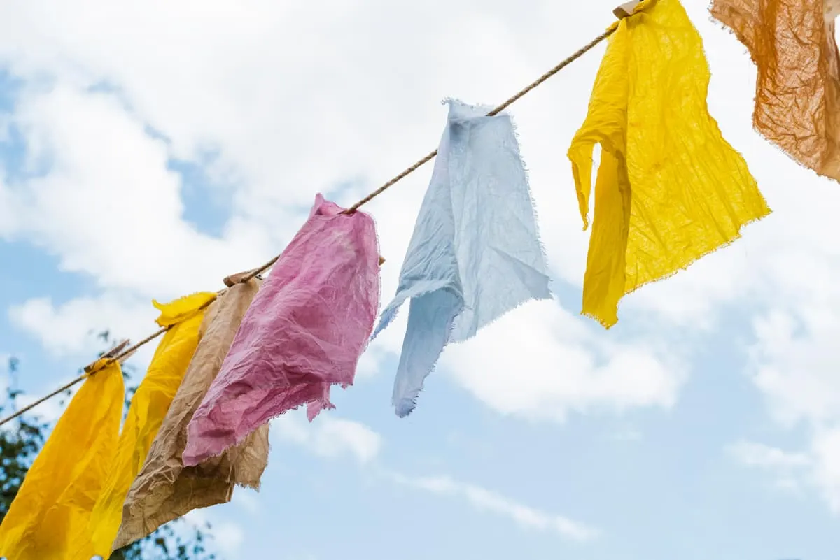 Imagen de ropa de colores azul, rosa, naranja y amarillo tendida en una cuerda de exterior. Fondo de cielo azul con nubes blancas.