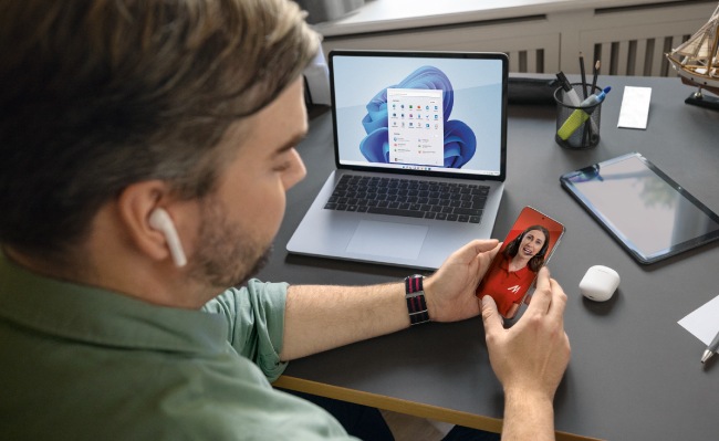 Un hombre con audífono usa un portátil con una foto en la pantalla y sostiene un móvil con otra foto sobre un escritorio.