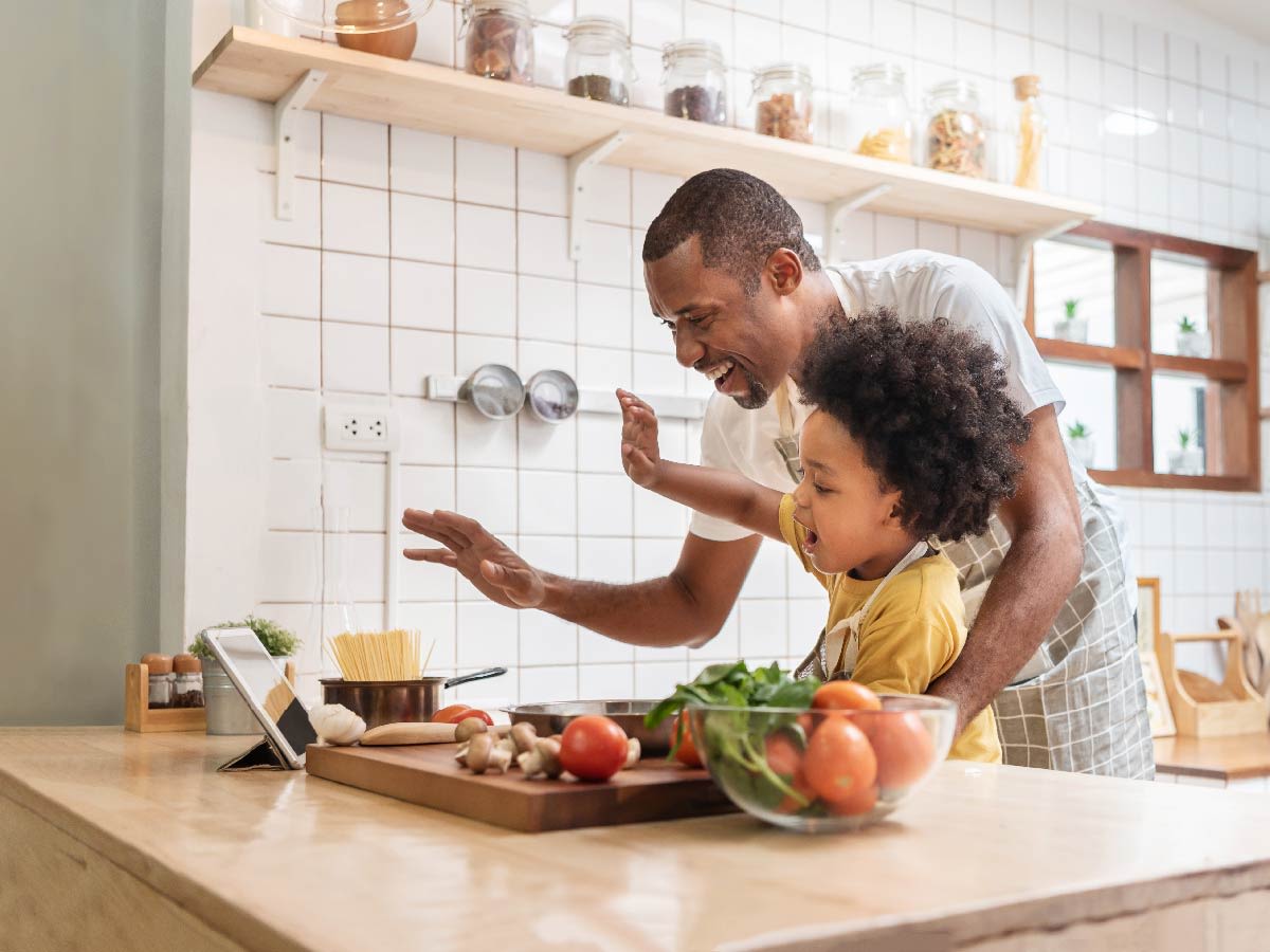 Persona adulta cocinando junto a un niño mientras consultan una tablet en la cocina.