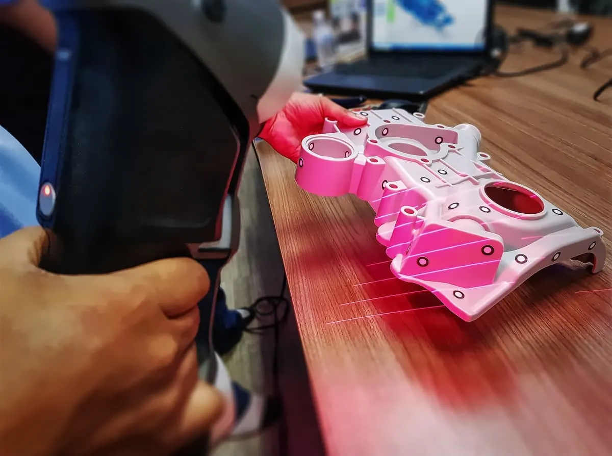 Mano usando un escáner 3D para digitalizar una pieza mecánica sobre una mesa.