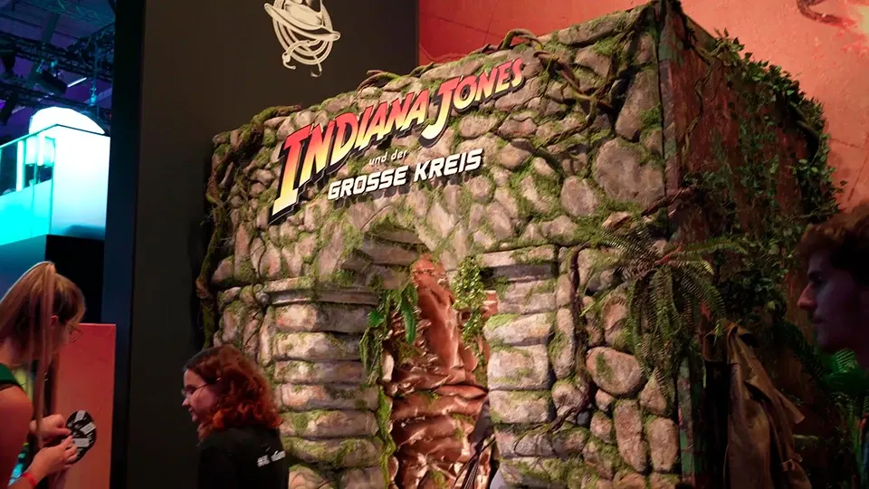 Decorado de Indiana Jones con texto en alemán y estructura de piedra cubierta de vegetación. Personas alrededor.