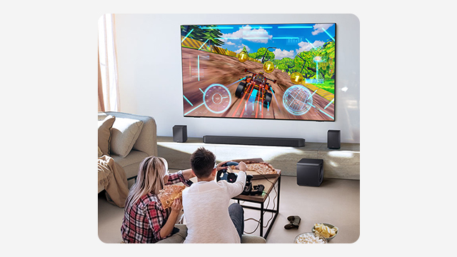 Pareja jugando videojuegos frente a una TV grande montada en la pared, con barra de sonido y bocinas en una sala cómoda.