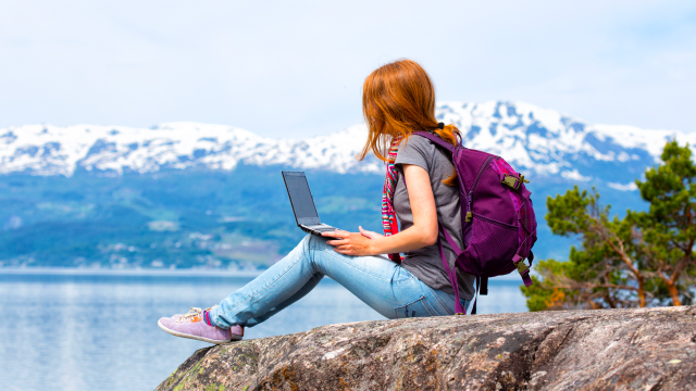Una persona con mochila morada está sentada en una roca con una laptop. Al fondo, se ve un lago y montañas nevadas.