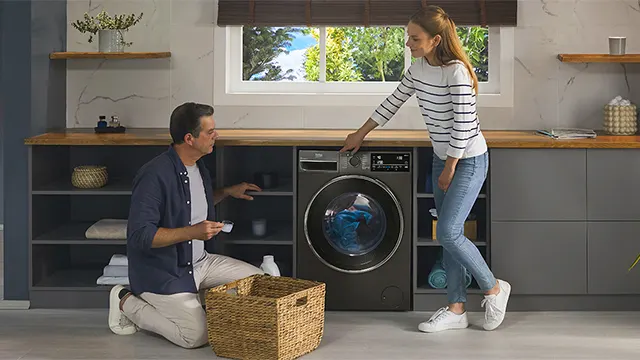 Un hombre retira la ropa de la lavadora y la coloca en un cesto. Una mujer está a su lado señalando la lavadora.