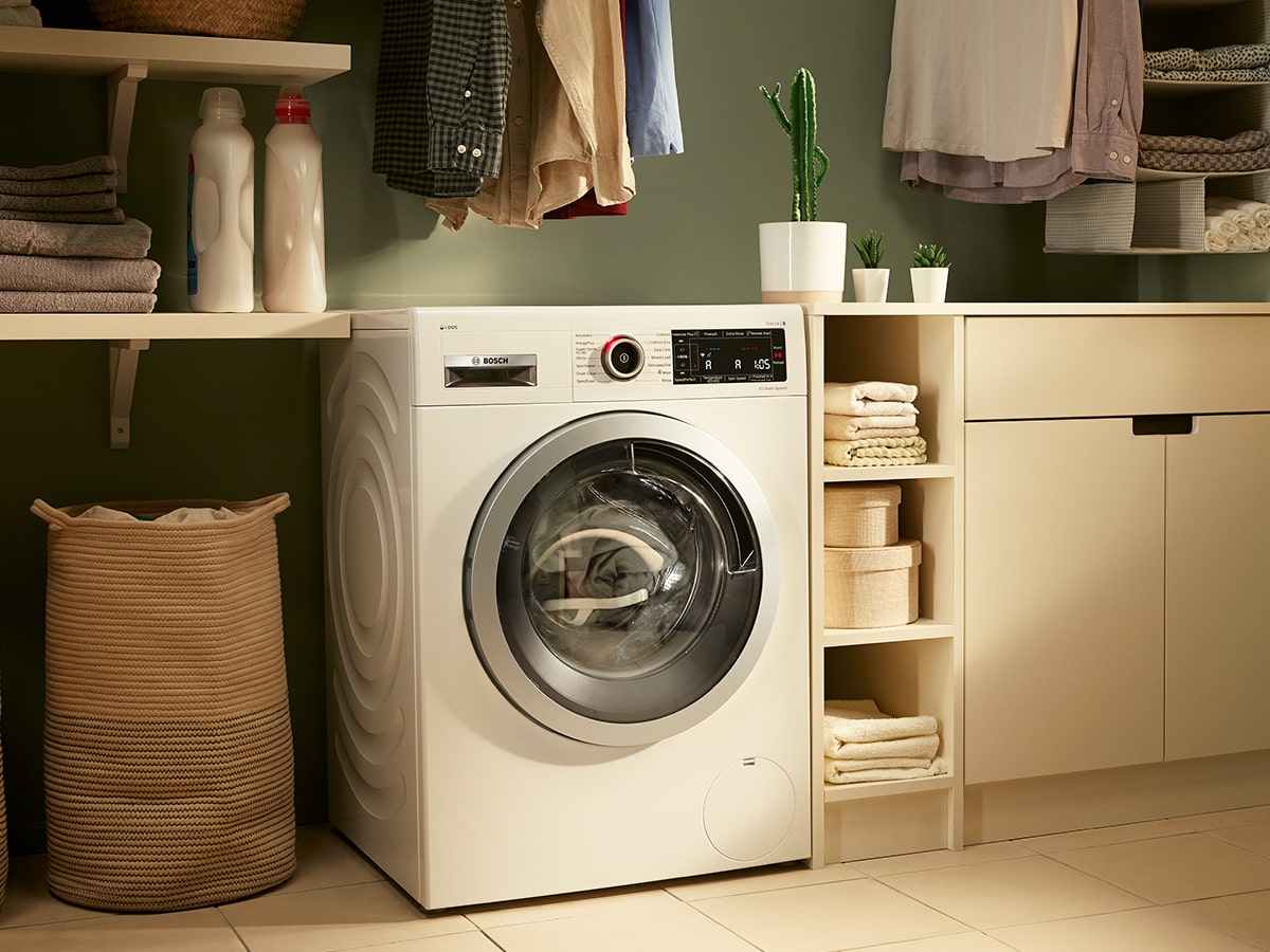 Lavadora blanca junto a cesto de ropa en cuarto con estantes y armarios. Hay toallas, plantas y detergente visibles.