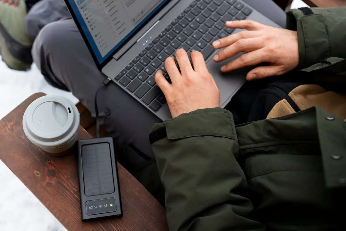 Persona con abrigo usando un portátil al aire libre, junto a un termo de café y un powerbank solar sobre la mesa.