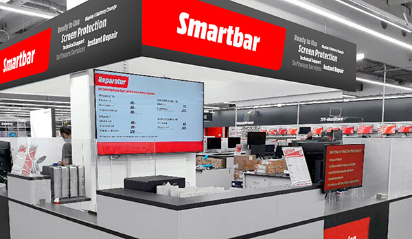 Mostrador Smartbar en tienda, con letreros rojos y negros. Pantalla muestra precios de reparación. Otros productos y mostradores visibles.