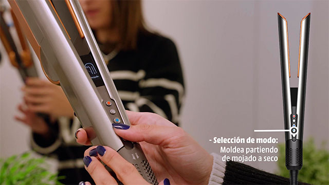 Plancha de pelo gris y dorada sostenida por una persona con las uñas pintadas de azul. Otra plancha se ve al fondo.