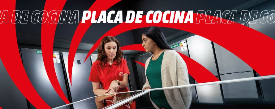 Esta imagen muestra a una empleada con uniforme rojo de MediaMarkt ayudando a una clienta a elegir entre diferentes modelos de placas de cocina en la tienda. El fondo presenta el característico diseño gráfico rojo y negro de la marca con el texto "PLACA DE COCINA" repetido