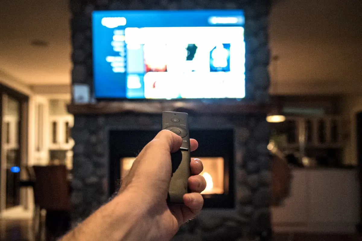 Mano apuntando un control remoto hacia un smart TV encendido sobre una chimenea en una sala de estar.
