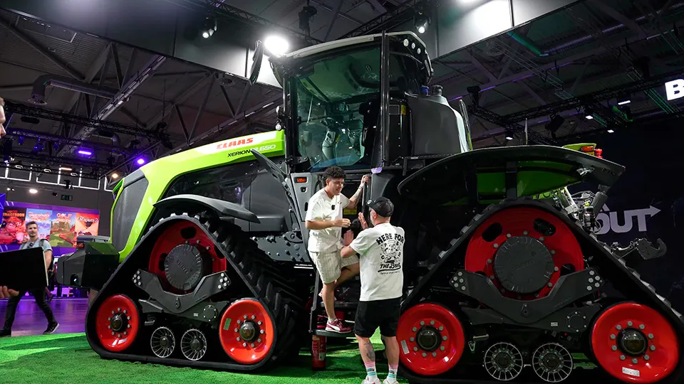 Dos personas cerca de un tractor verde y gris con ruedas rojas en un evento. El piso es verde.