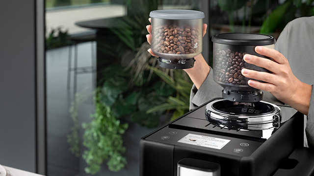 Persona llenando una máquina de café negra con granos de café desde un recipiente transparente. Hay plantas en el fondo.