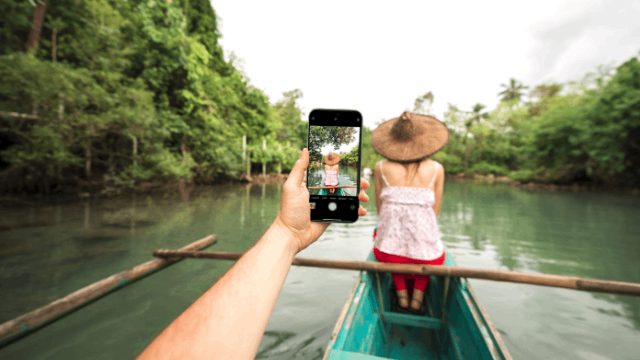 En un río bordeado de árboles, una persona toma una foto con un móvil a una mujer con sombrero sentada en un bote.