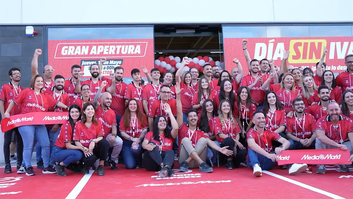 Grupo de personas con camisetas rojas en la apertura de una tienda. Globos rojos y blancos adornan la entrada. Carteles de "Gran Apertura" y "Día sin IVA".