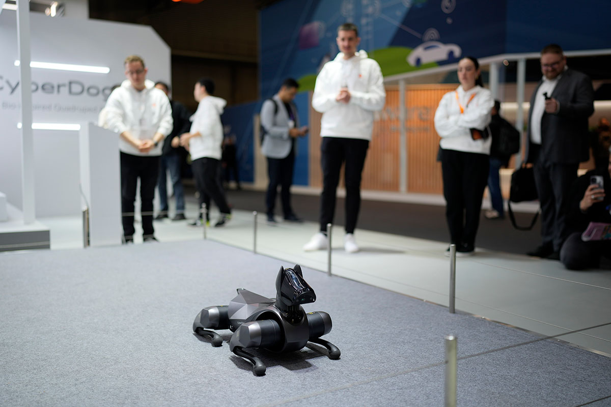 Un perro robótico negro en exhibición sobre una alfombra gris, con gente de pie al fondo.