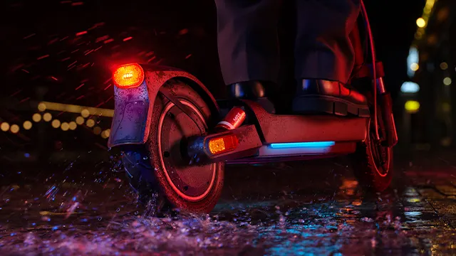Rueda trasera del Xiaomi Electric Scooter 5 Max salpicando agua en una calle mojada durante la noche, con luces encendidas y reflejos urbanos.
