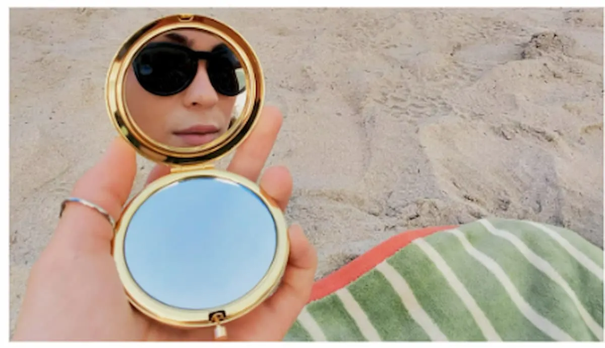 Mujer con gafas de sol Ray-Ban Meta reflejada en un espejo de bolsillo en la playa