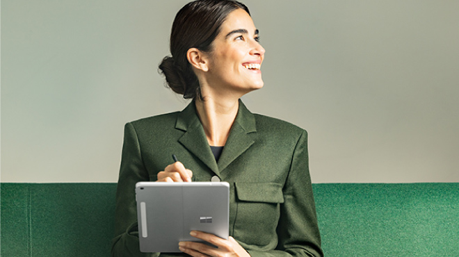 Una persona sostiene una tableta frente a un edificio con plantas anaranjadas. Lleva una camisa blanca debajo de un chaleco azul marino.