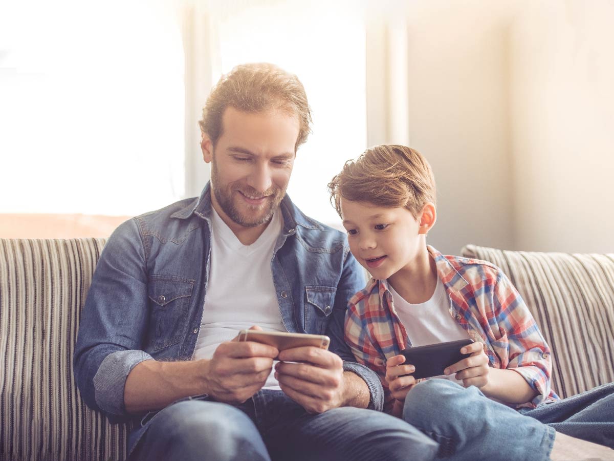 Persona adulta y niño sentados en el sofá, cada uno usando un dispositivo móvil.