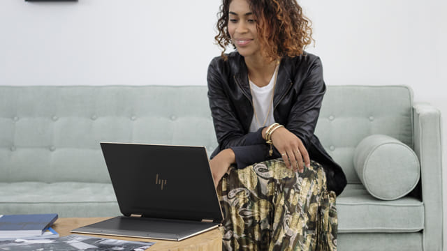 Una mujer está sentada en un sofá gris con un portátil negro en una mesa baja. Ella lleva una chaqueta negra y una falda estampada.