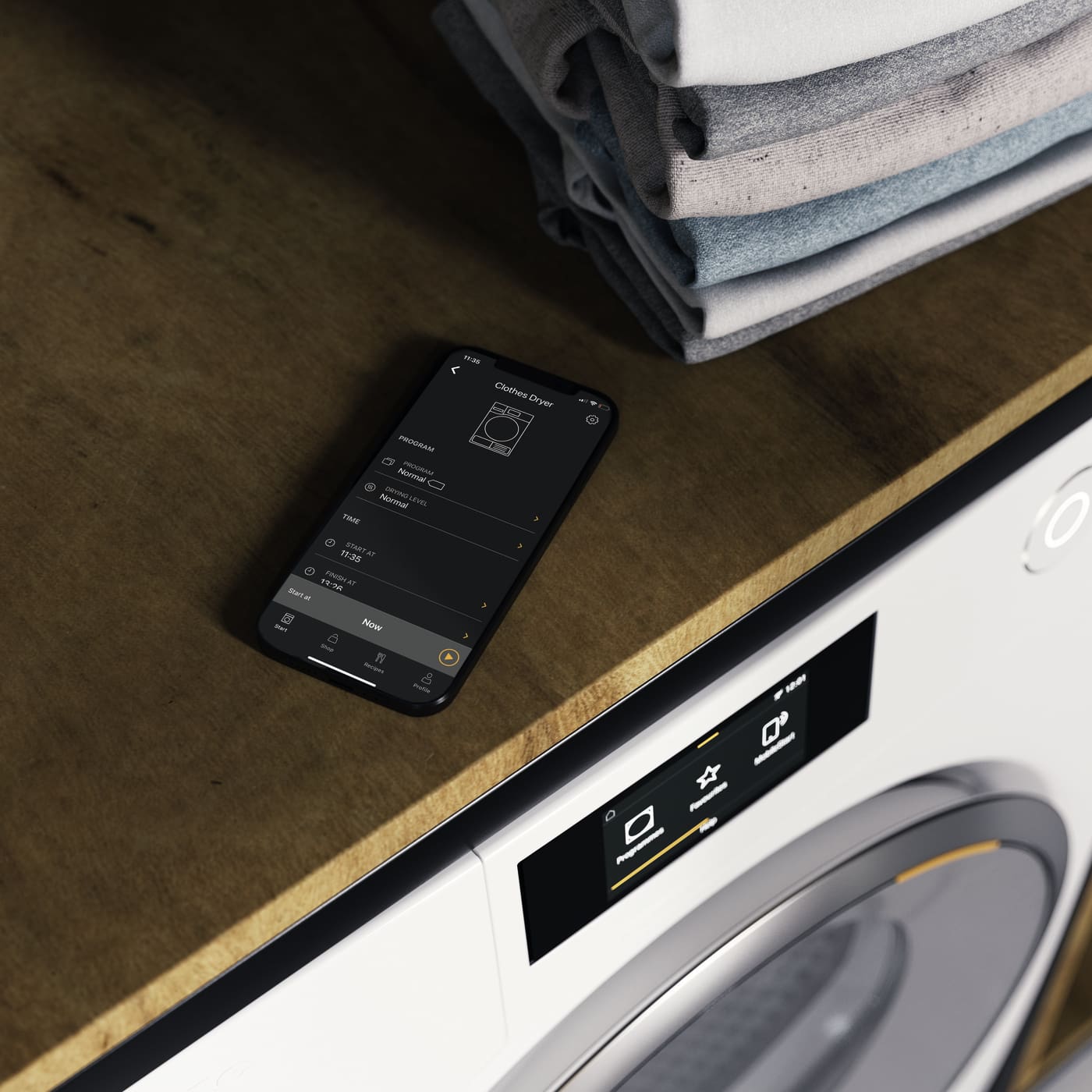 Un teléfono muestra controles de secadora junto a una pila de ropa y una secadora blanca. La pantalla es negra con texto blanco.