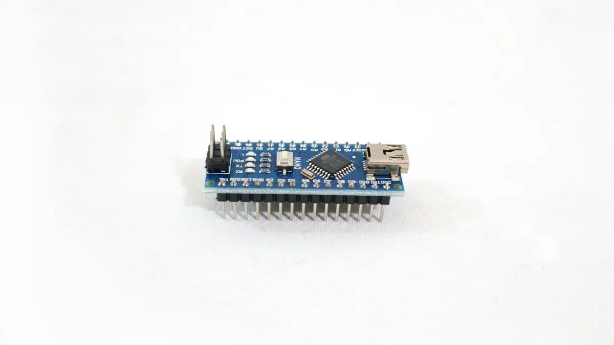 Placa de circuito azul con un chip cuadrado negro, clavijas plateadas y un puerto USB sobre un fondo blanco.