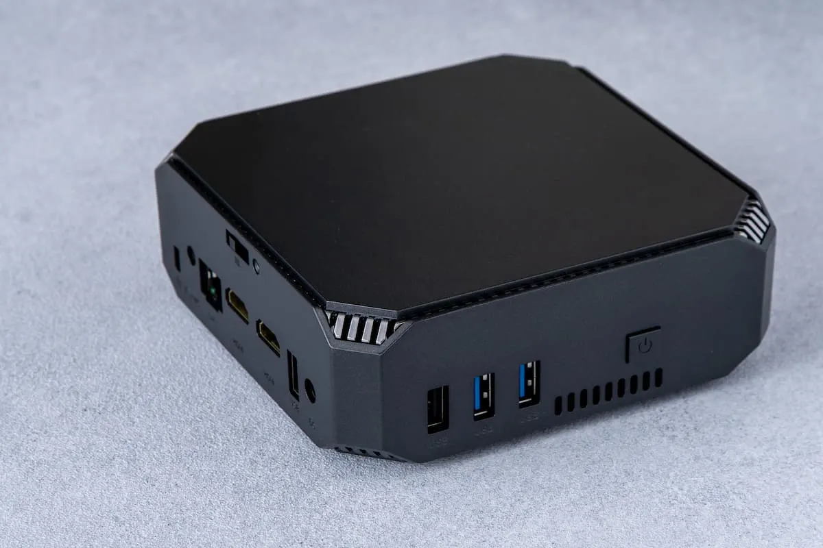 Mini PC negro rectangular sobre una superficie gris, mostrando varios puertos USB y de vídeo en el frontal.