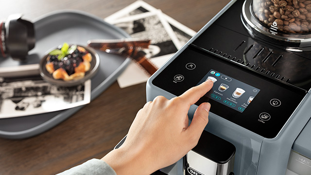 Una mano toca la pantalla de una máquina de café gris y negra. En una bandeja gris hay fruta, fotos y gafas.