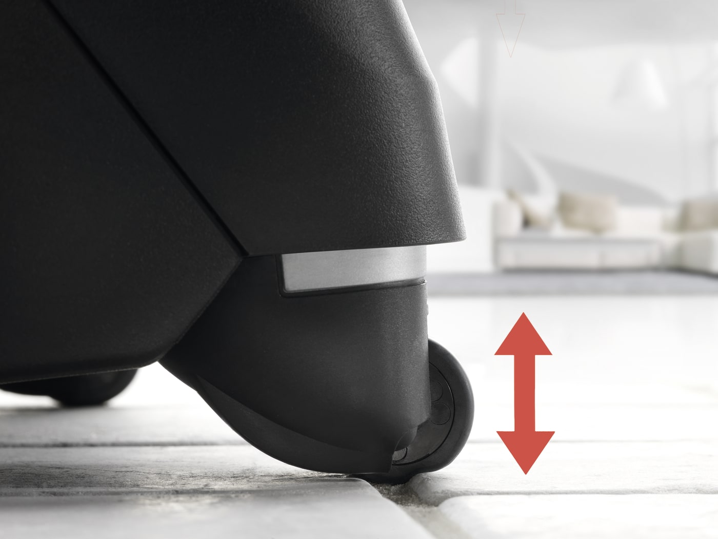 Primer plano de la rueda de una silla negra sobre un suelo de baldosas. Una flecha roja señala hacia arriba y abajo.