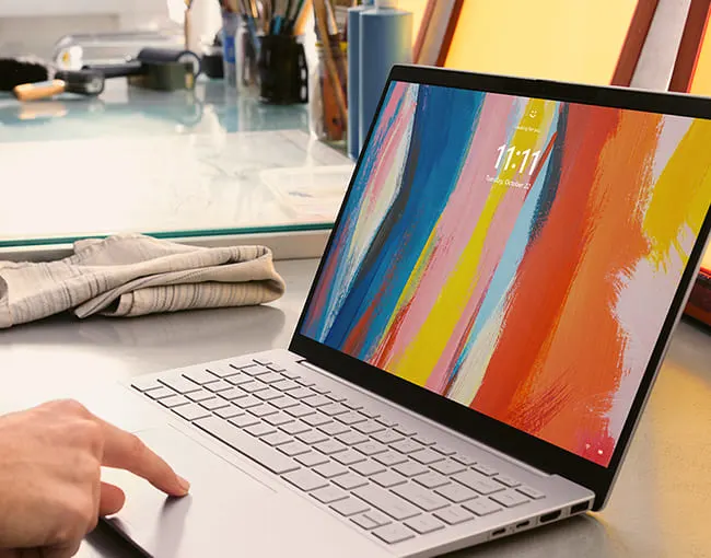 Ordenador portátil abierto mostrando pantalla de colores con la hora 11:11. Una mano toca el panel táctil. Al fondo, artículos de arte.