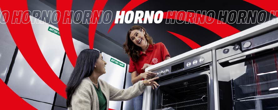 Una empleada de tienda con polo rojo explica las funciones de un horno a una clienta; la clienta señala los mandos mientras examinan el electrodoméstico.
