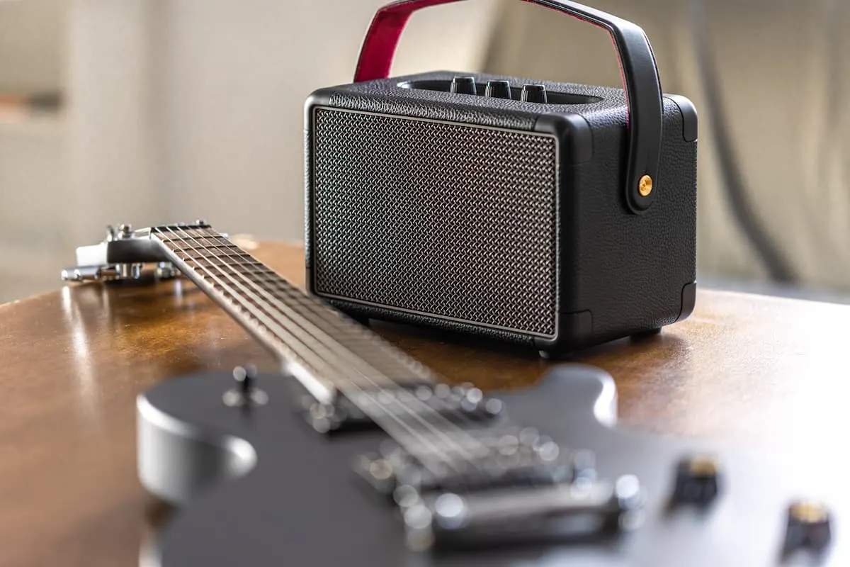 Altavoz portátil negro con asa junto a una guitarra eléctrica sobre una mesa de madera.